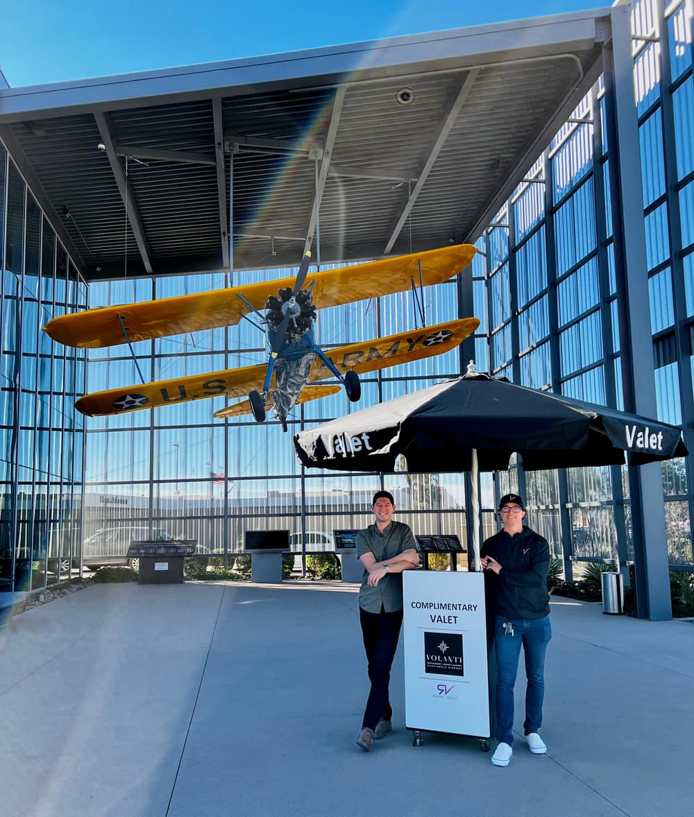 Two Royal Valet attendants at a complimentary valet stand under a vintage biplane at Volanti